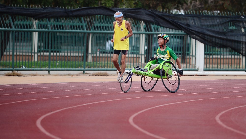 En gammal man som springer bredvid en kille som är funktionshindrad som cyklar